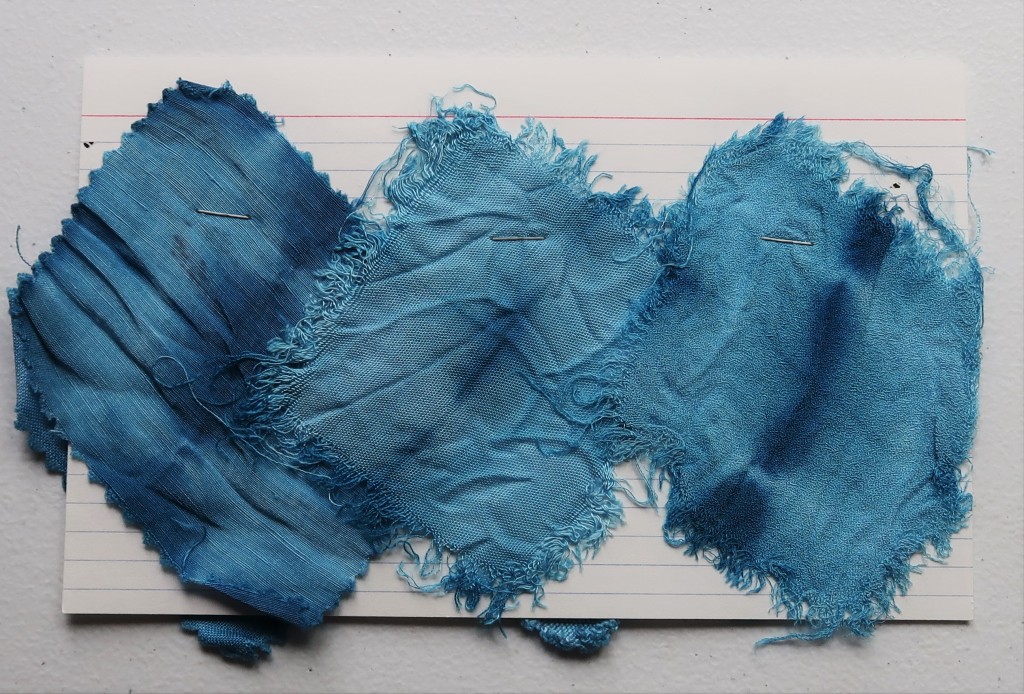 An index card on a white table. Three fabric swatches are taped to the card with black and white washi tape. All three are light blue with dark blue fold lines. The left and middle swatches are thin, easily wrinkled fabrics.