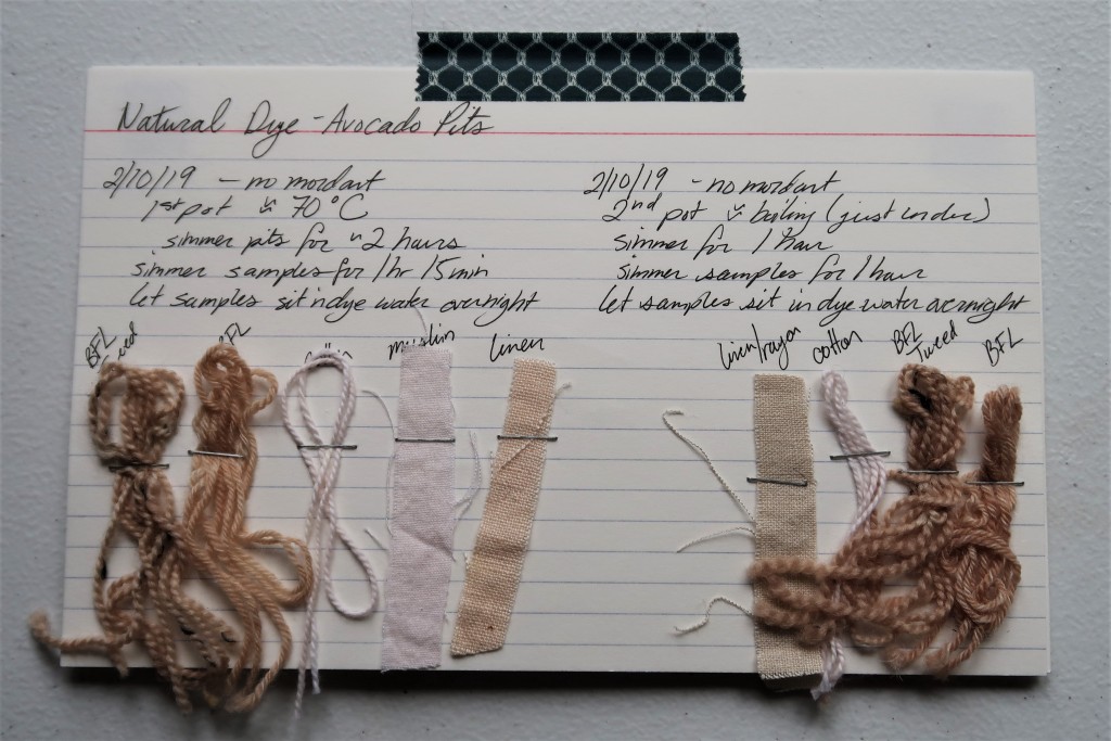 An index card on a white table. There is information for the first and second avocado pit dye baths written in black ink that is detailed on this web page. 

Beneath the first dye bath are 3 yarn samples and 2 fabric samples. The BFL yarn samples are a pinkish tan color. The cotton yarn is a very light pink. The muslin fabric sample is just off white pink. The linen fabric sample is a light pinkish tan color.

There are 3 yarn samples and 1 fabric sample stapled beneath the second dye bath information. The fabric sample is a light greyish pink. The cotton yarn is a similar color with less of a grey tone. The two BFL yarns are a grey mauve.