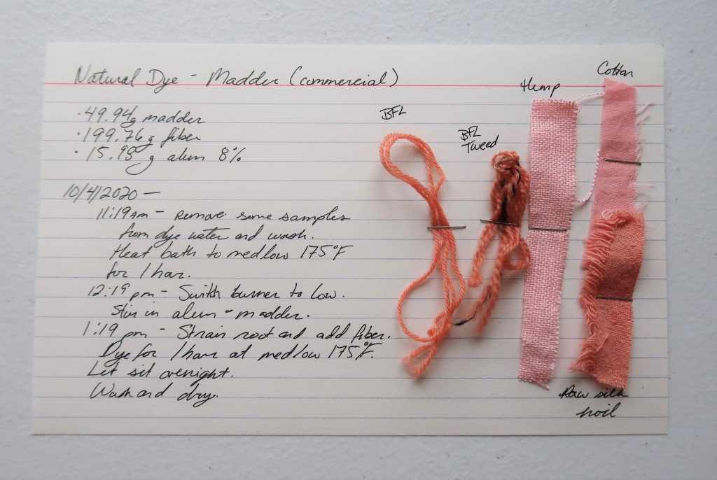 An index card on a white table. On the left, the information from this post is hand written in black ink.

There are 5 samples stapled to the left of the card. The BFL yarn is a salmon color. The BFL Tweed is a light red or dark salmon color. The hemp fabric sample is pink, The cotton sample is a pink leaning in the red orange direction. The raw silk noil fabric sample is a light red.