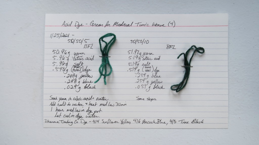 An index card on a white table. Two columns of information from this post are handwritten in black ink. At the top of the left column is a BFL yarn sample in a medium green. At the top of the right column is a BFL yarn sample is a dark green.